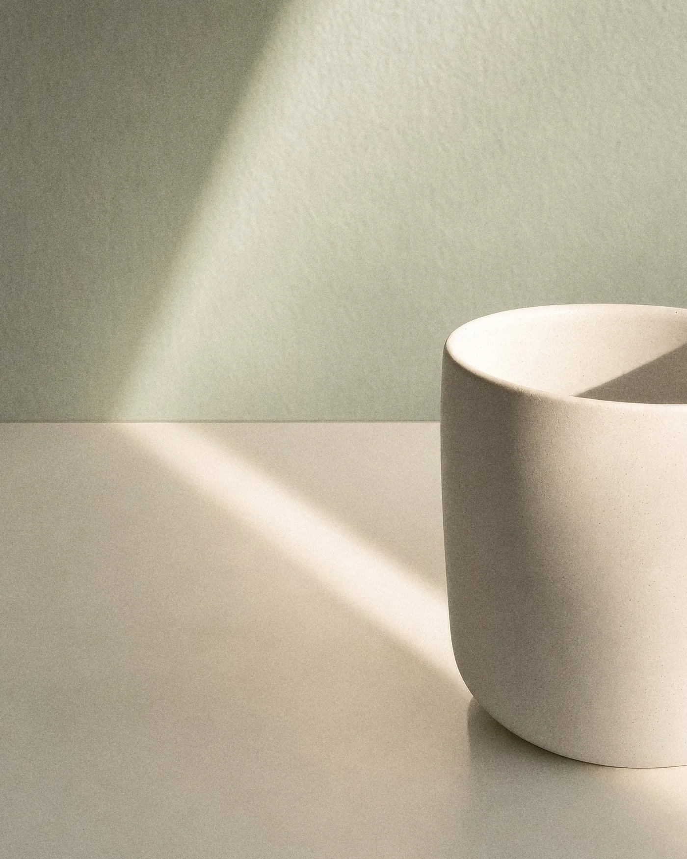 Ein schlichter weißer Becher im Streifen Sonnenlicht vor einer salbeigrünen Wand, mit KI erzeugt