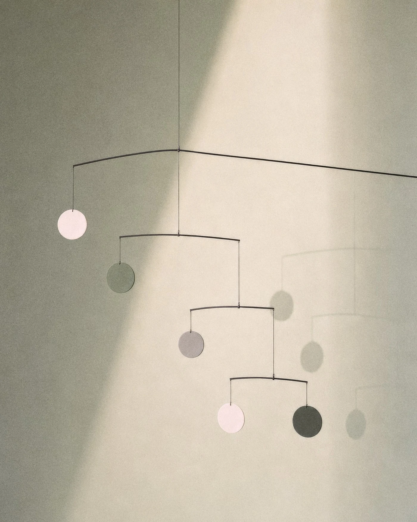Ein schlichtes Mobile aus dünnen schwarzen Drähten und runden Scheiben hängt im Sonnenlicht vor einer salbeigrünen Wand, sein Schatten daneben, mit KI erzeugt