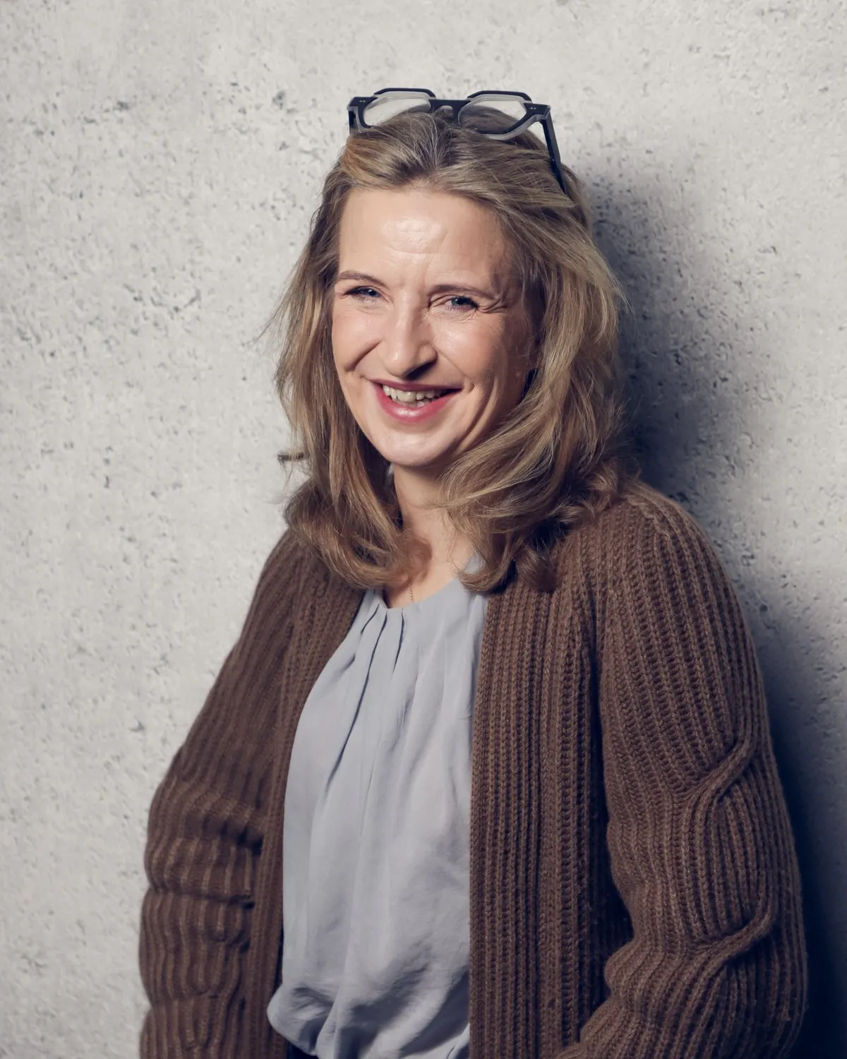 Katrin Funke, lachend, im braunen Cardigan vor einer Betonwand